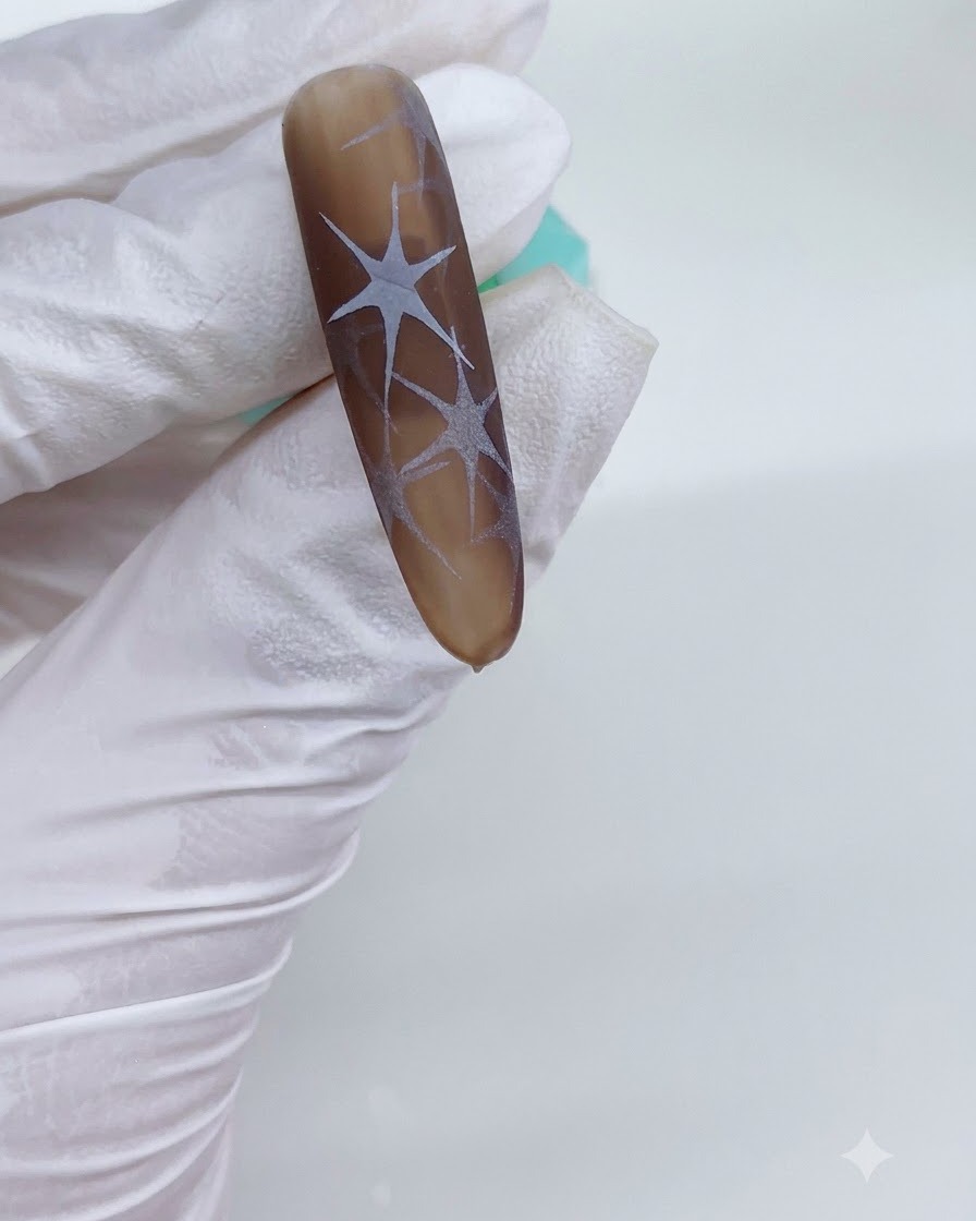 Une main gantée tenant un ongle aux tons bruns sous une lampe UV avec des motifs détoiles blanches catalysés visibles.