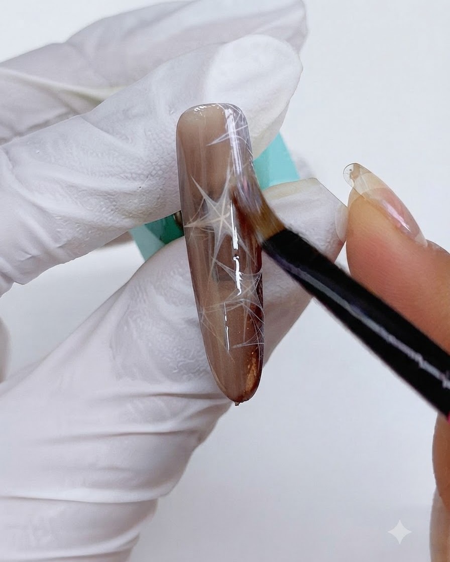 Un pinceau fin appliquant du gel cat eye sur longle brun décoré.