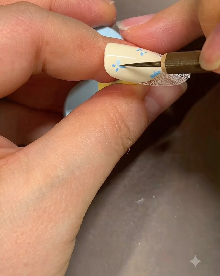 Un pinceau de nail art fin ajoutant des accents bleus détaillés au centre dune fleur peinte sur un ongle beige.