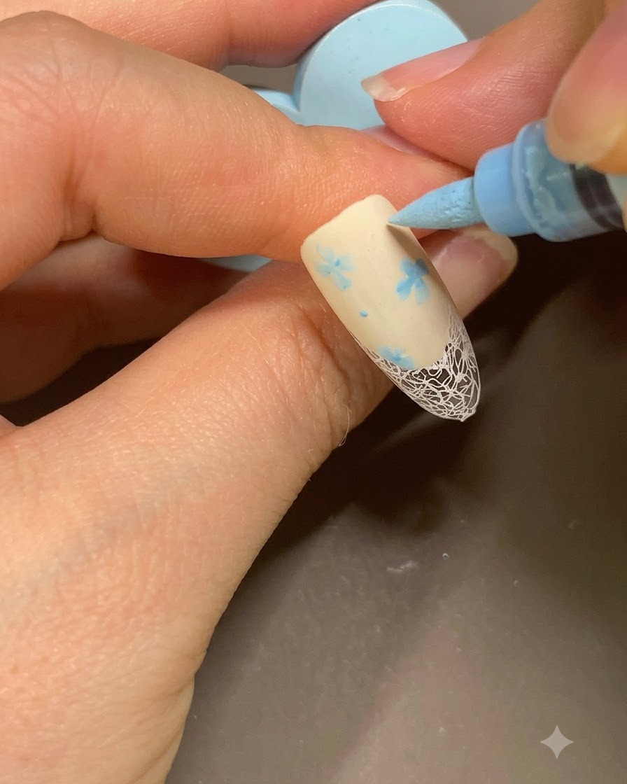 Un marqueur de nail art bleu ajoutant de petits points finaux à côté dun motif floral sur une french manucure beige.