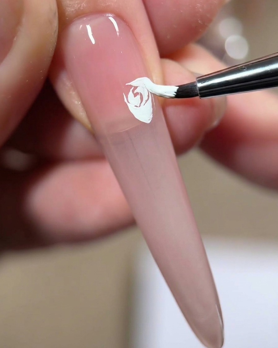 Un pinceau à ongle pressant et étirant du gel blanc pour ajouter un quatrième pétale superposé à une rose blanche peinte sur un ongle rose.
