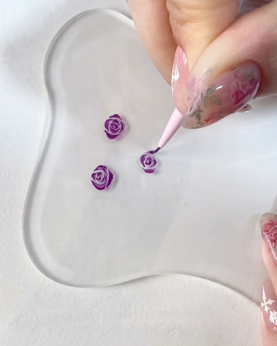 Un gros plan dune main tenant un pinceau de détail fin, peignant méticuleusement des accents violet foncé sur un petit motif de rose 3D sur une palette en verre transparente.