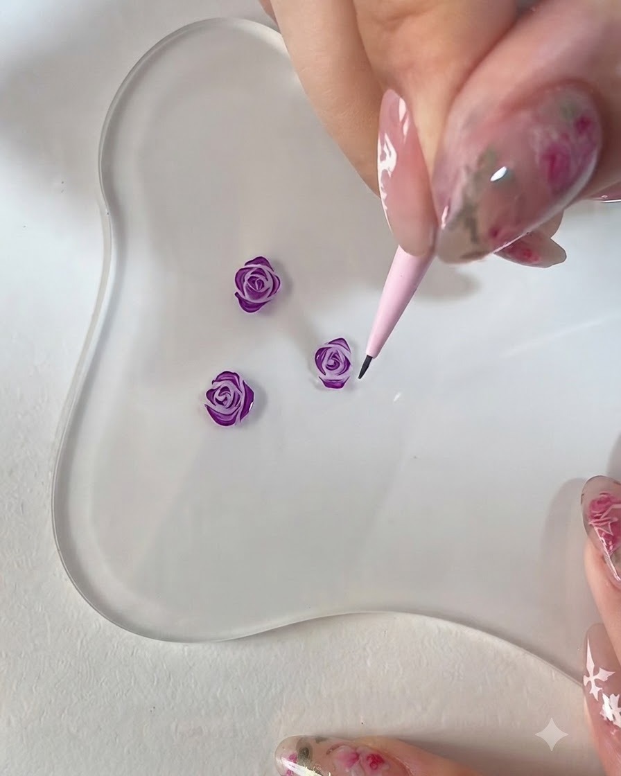 Un gros plan dune main utilisant un pinceau de détail rose pour affiner les bords dune rose violette peinte en 3D sur une palette transparente.