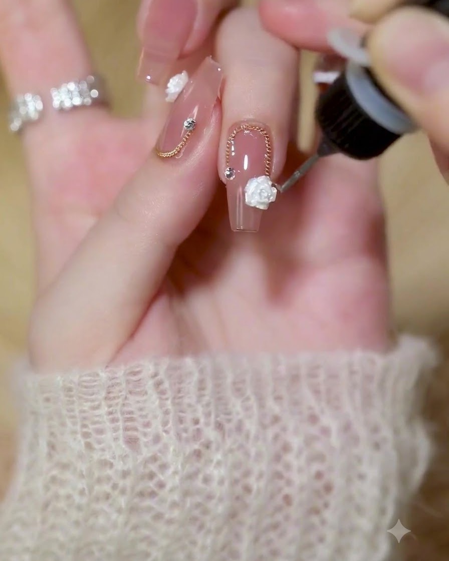 Un pinceau de détail fin appliquant du gel de construction transparent autour des bords dune rose blanche 3D et dune chaîne dorée sur un ongle vernis.