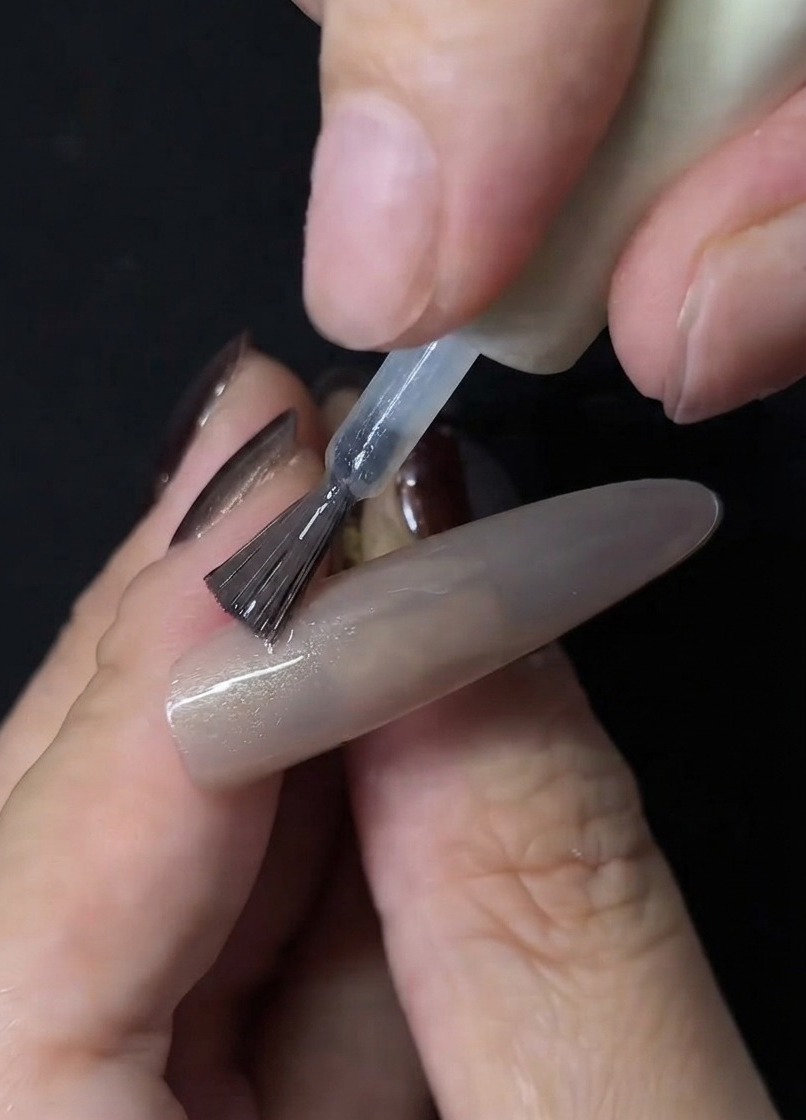 Un pinceau à ongles appliquant du gel cat-eye transparent sur la surface dune extension dongle nude.