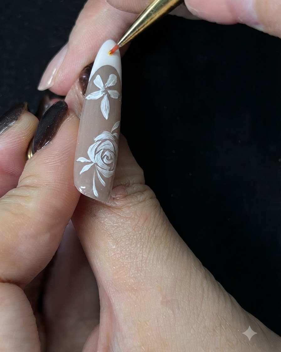 Gros plan dune main utilisant un pinceau fin pour peindre de petites vignes blanches autour dune rose peinte à la main sur un ongle en gel nude.