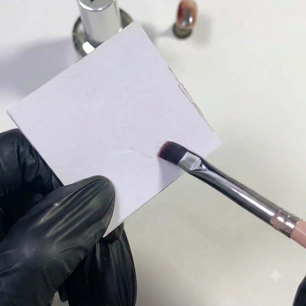 Un pinceau à ongles plat tamponnant du gel de base transparent sur une surface de palette blanche.