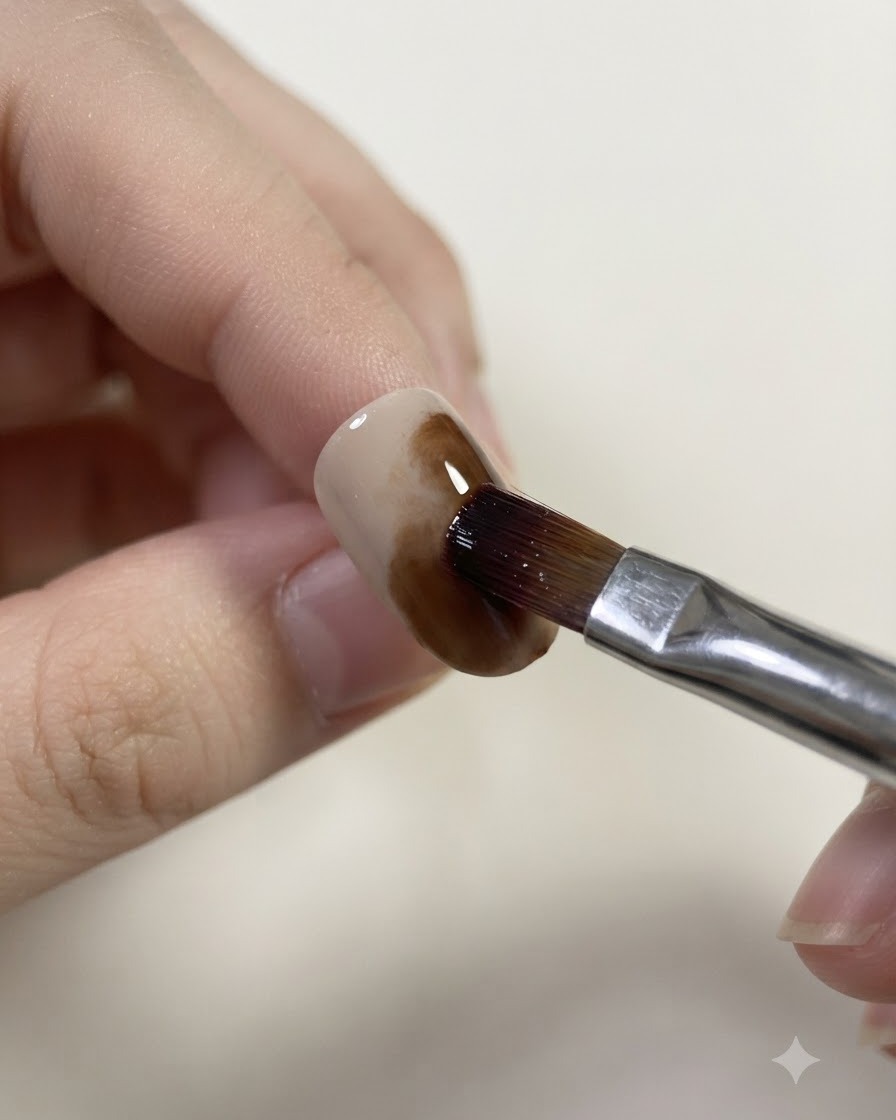 Un pinceau humidifié mélangeant et adoucissant les bords du gel caramel sur un ongle.
