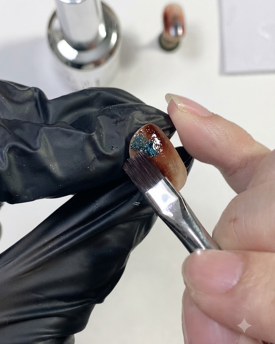 Un pinceau à ongles plaçant une petite touche de gel œil-de-chat magnétique bleu scintillant sur le côté de longle ambre.