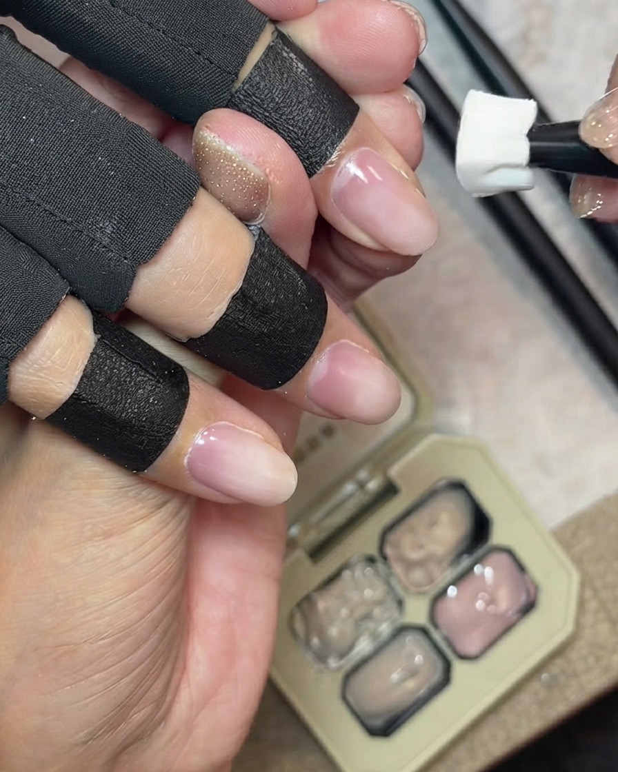 Une éponge tamponnant un gel couvrant nude laiteux sur le bord libre naturel dun ongle, avec une palette de gel en arrière-plan.
