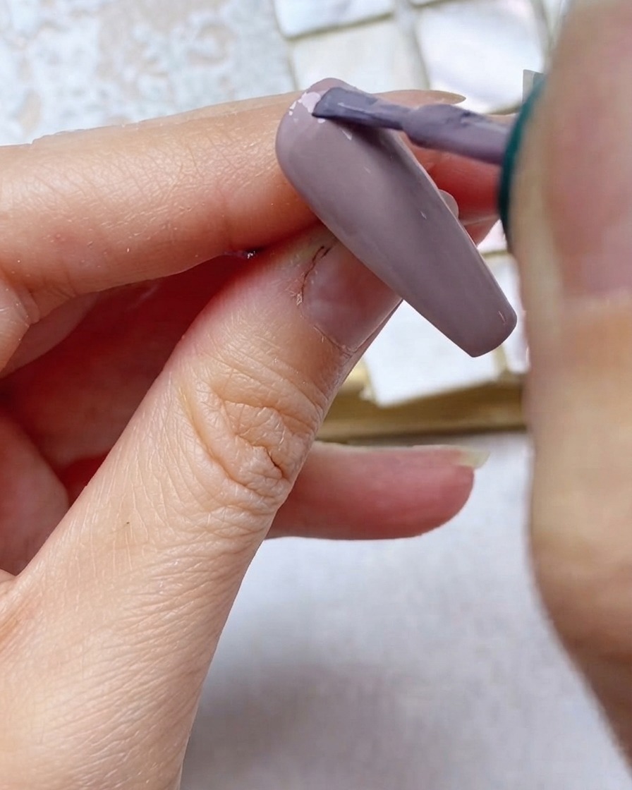 Un pinceau appliquant du vernis gel gris-violet sur le tiers supérieur de longle près de la cuticule.