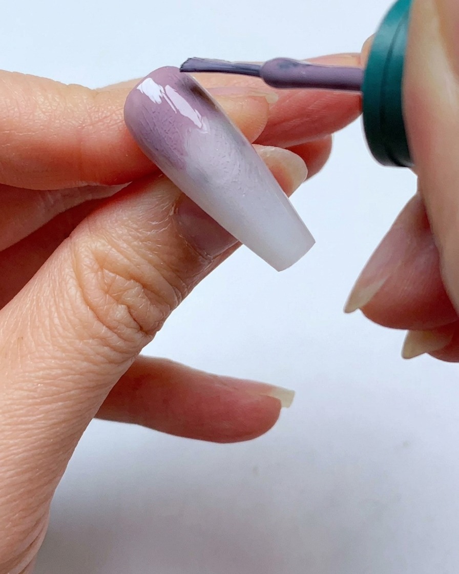Un pinceau à vernis gel appliquant une deuxième couche de gel gris-violet près de la cuticule dun ongle.