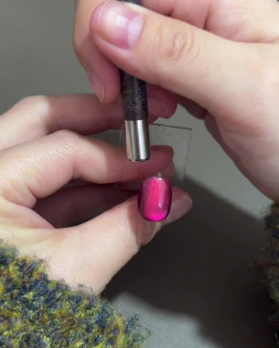 Une main tenant un aimant cylindrique au-dessus du bord supérieur dun ongle en gel violet pour pousser le reflet magnétique vers le bas.