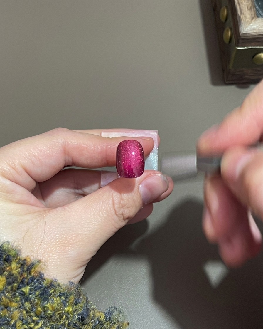 Une main tenant un petit aimant cylindrique près dun ongle en gel cat-eye violet.
