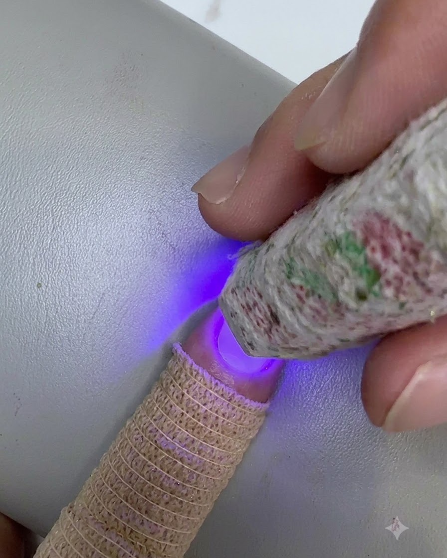 Une mini lampe UV/LED à motifs tenue au-dessus du bout dun ongle, catalysant une couche estompée de gel transparent.