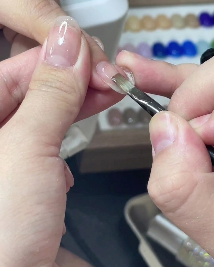 Un pinceau fin appliquant un vernis gel rose diaphane uniformément sur un ongle.
