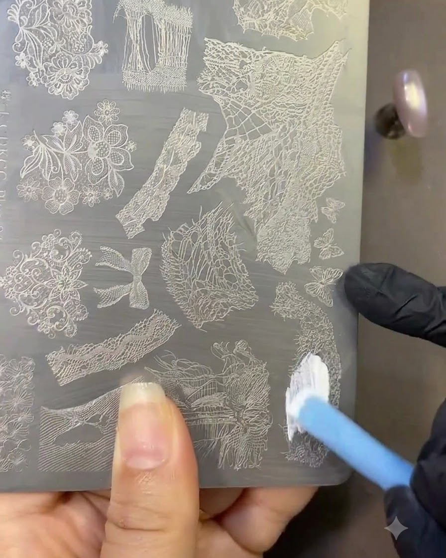 Un pinceau appliquant du vernis de stamping blanc sur une plaque métallique gravée de motifs de dentelle.