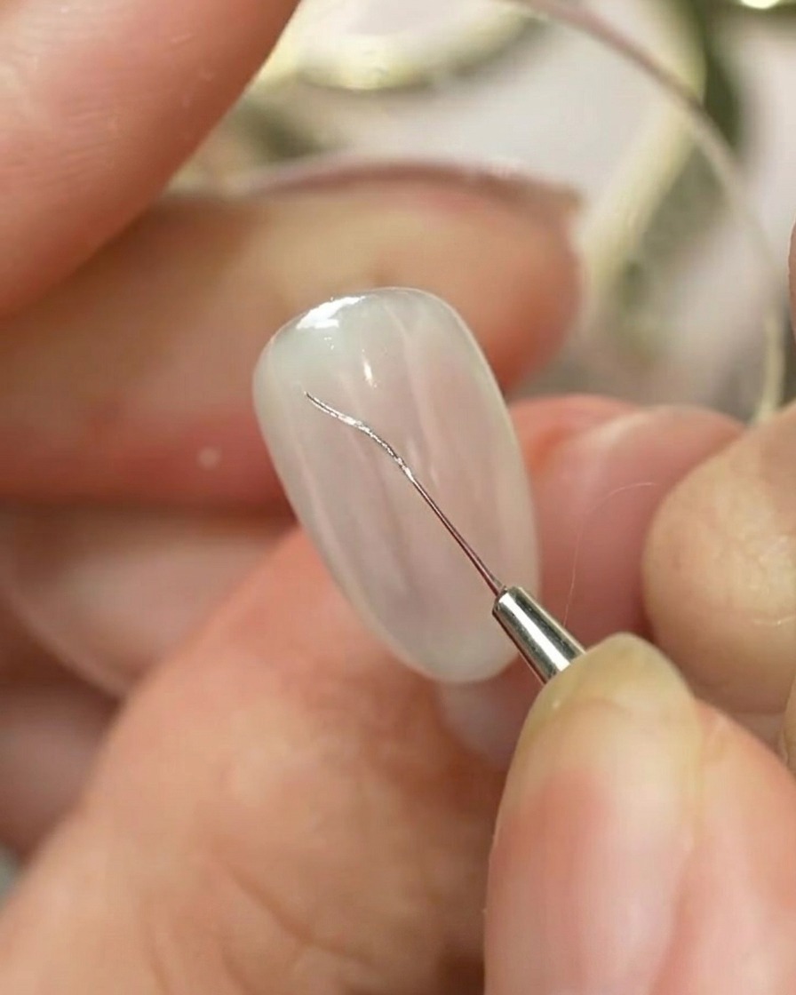 Un pinceau fin pour nail art dessinant un contour argenté de nœud sur un onglet manucuré de couleur claire.
