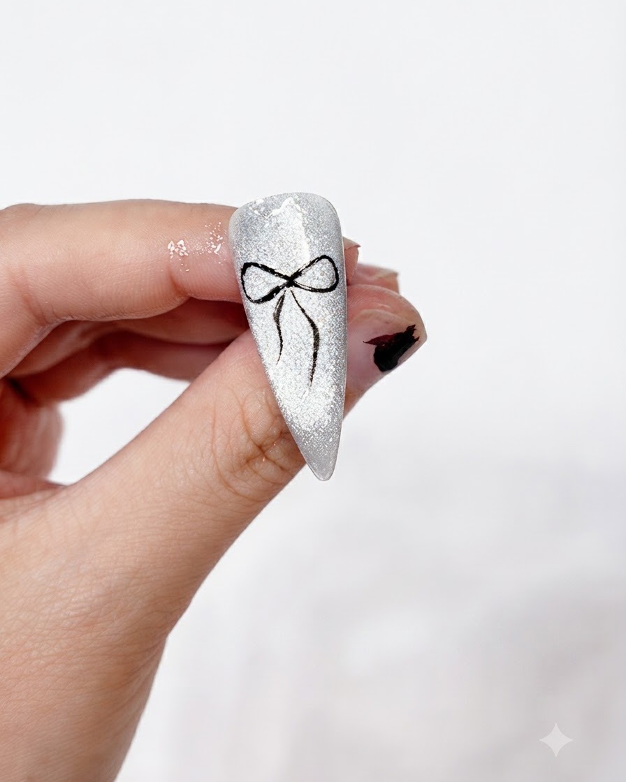 Un nail art de nœud noir terminé, proprement peint et fixé sur un fond cat-eye argenté étincelant, tenu entre les doigts.