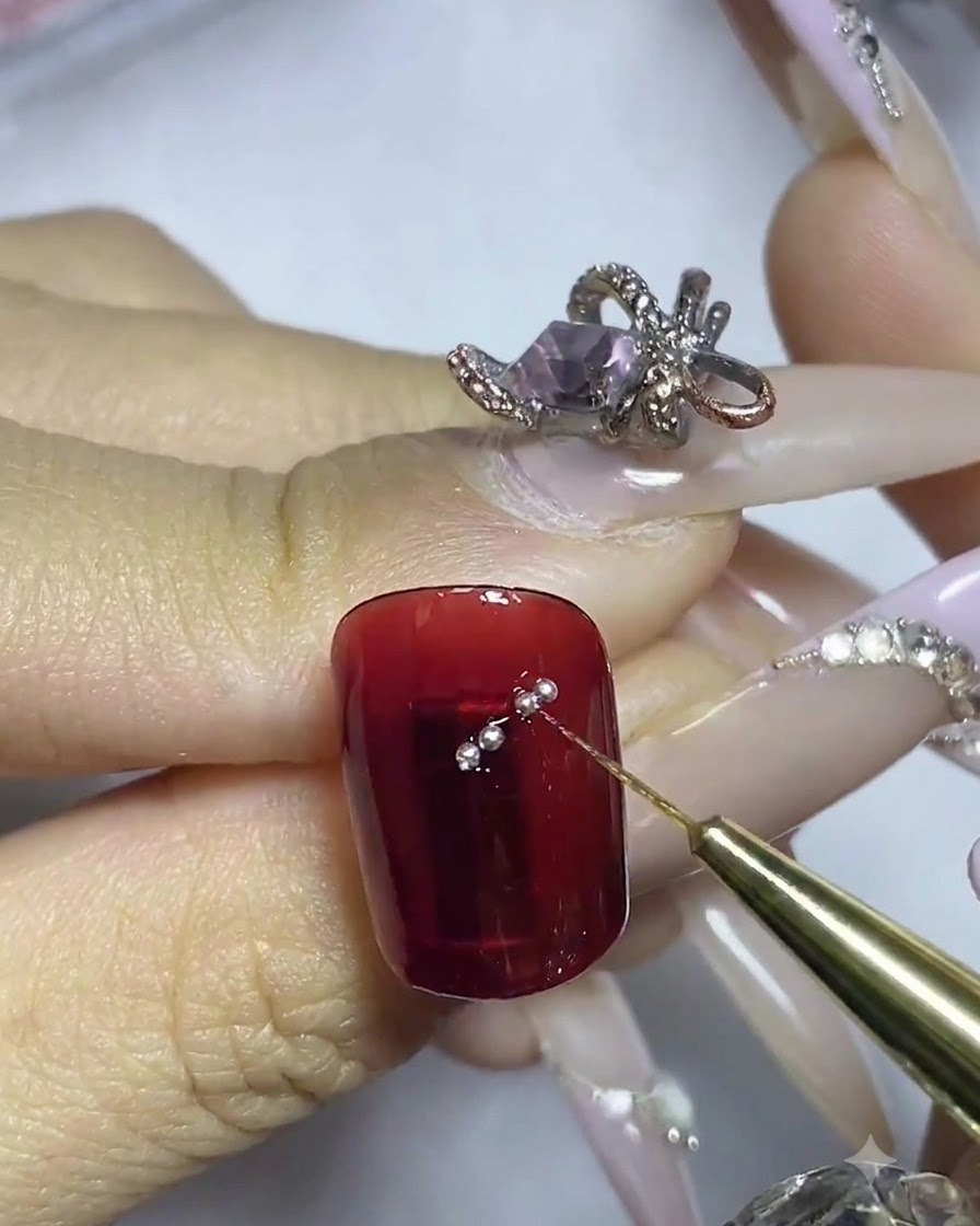 Un pinceau fin plaquant de petites billes dacier argentées le long du tracé adhésif sur un ongle rouge.