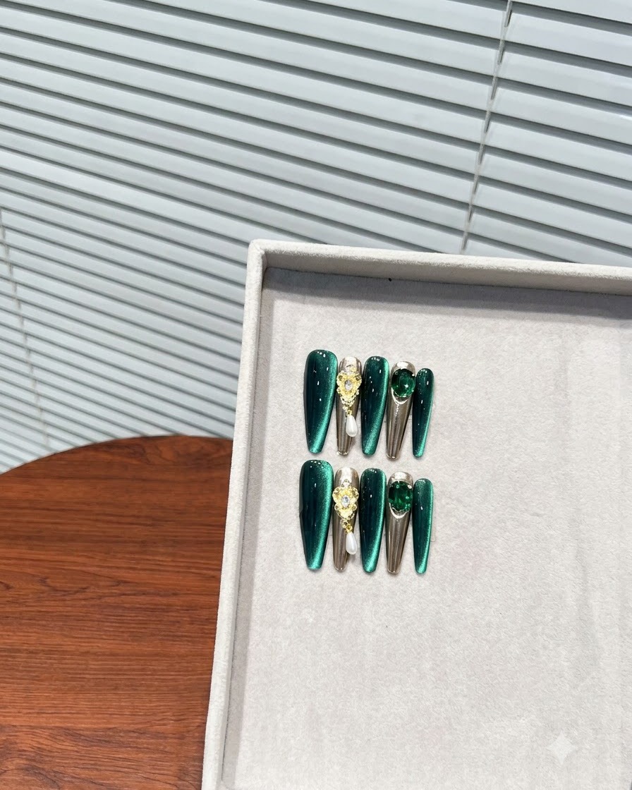 Une vue rapprochée en biais dune boîte de rangement contenant un jeu complet de faux-ongles longs en amande vert foncé avec des accents métalliques et de strass.