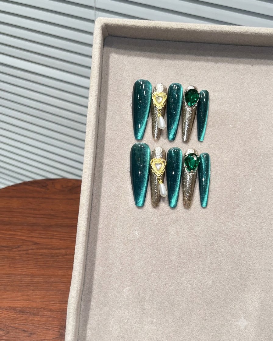Un plan rapproché axé sur la section gauche des faux-ongles vert émeraude présentant des dégradés cat-eye et des charms en forme de cœur ornés de perles suspendues.