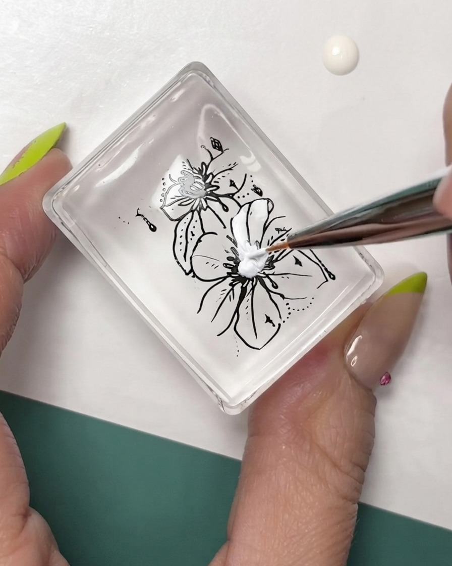Un pinceau fin pour nail art ajoutant des détails blancs sur un estampage floral à contour noir sur un tampon transparent.