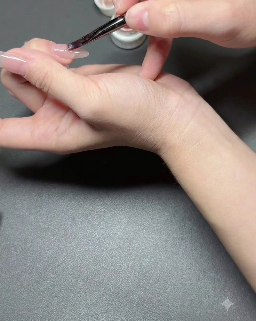 Un pinceau à ongles appliquant une fine couche de vernis nude semi-transparent sur un ongle.