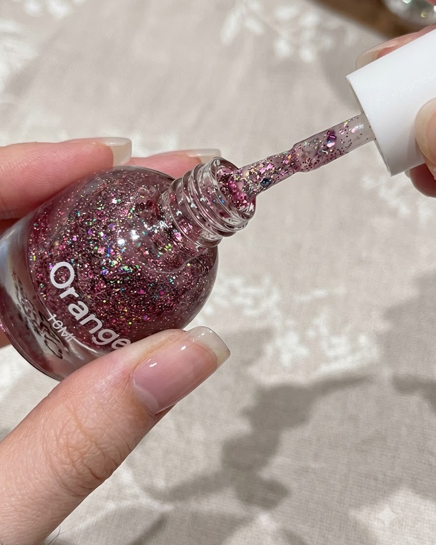 Un pinceau appliquant une fine couche de gel pailleté rose sur la moitié supérieure dun ongle naturel.