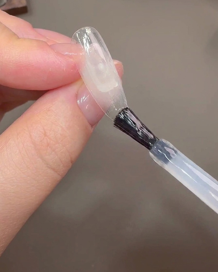 Un pinceau à ongles appliquant du gel de construction sur un ongle transparent pour couvrir la ligne de croissance.