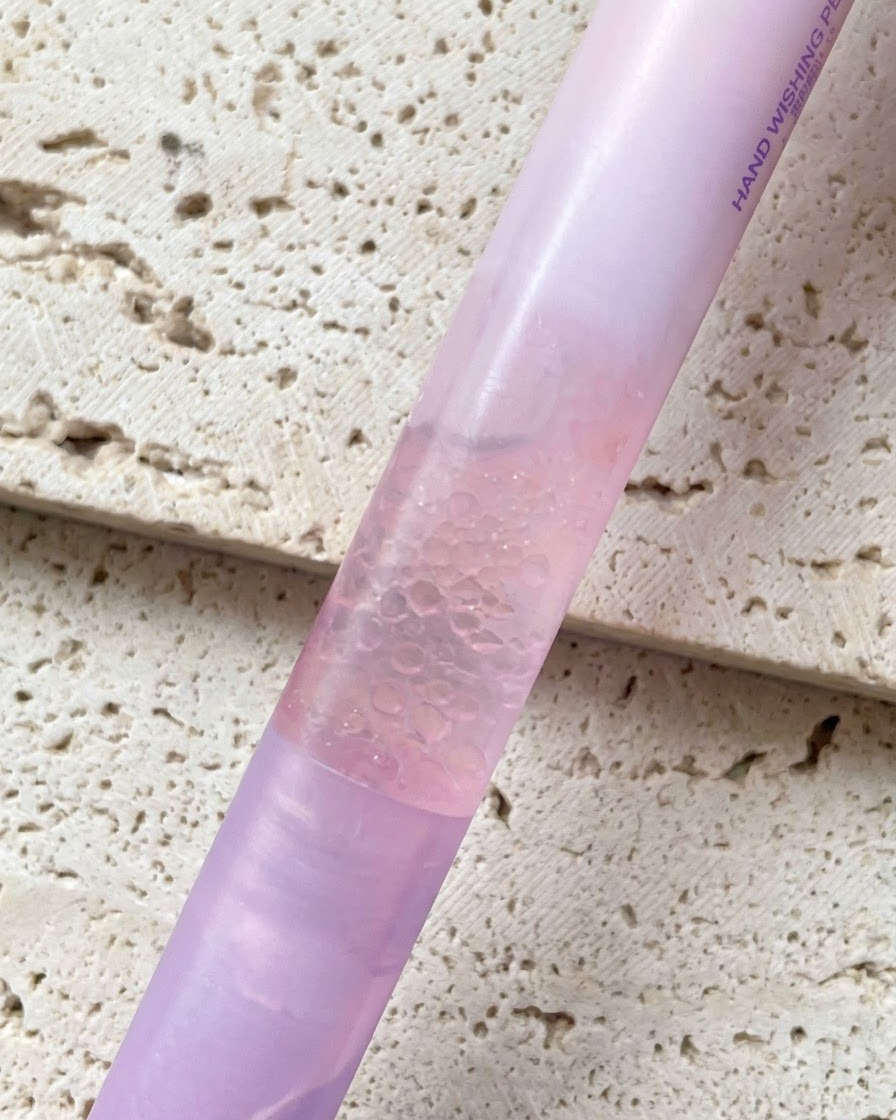 Un stylo huile pour cuticules à lessence de perle au dégradé rose tenu devant un fond texturé.