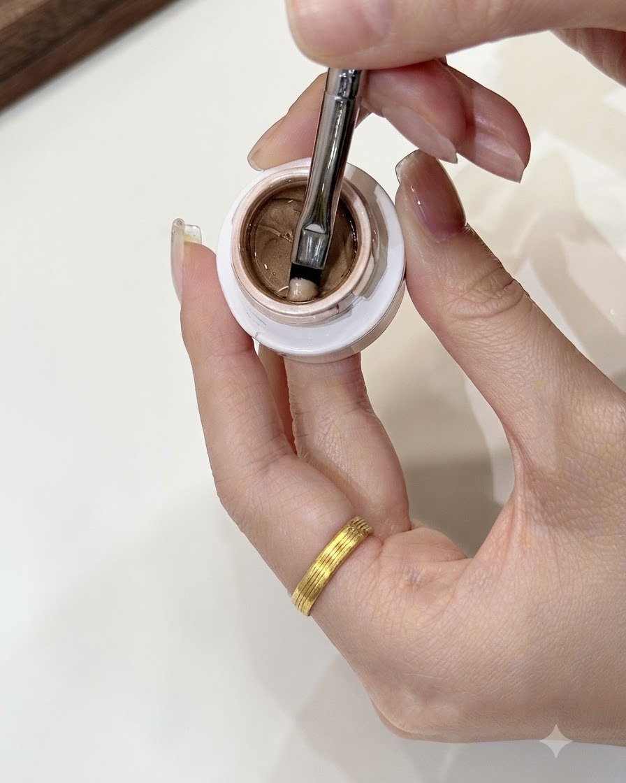Un pinceau de nail art prélevant une petite portion de gel or champagne de la taille dun grain de haricot mungo dans un pot ouvert.
