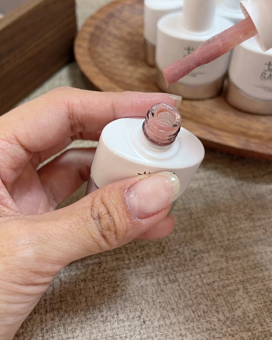 Un pinceau applicateur avec du vernis gel nude rose pêche miellé tenu au-dessus dun flacon ouvert.