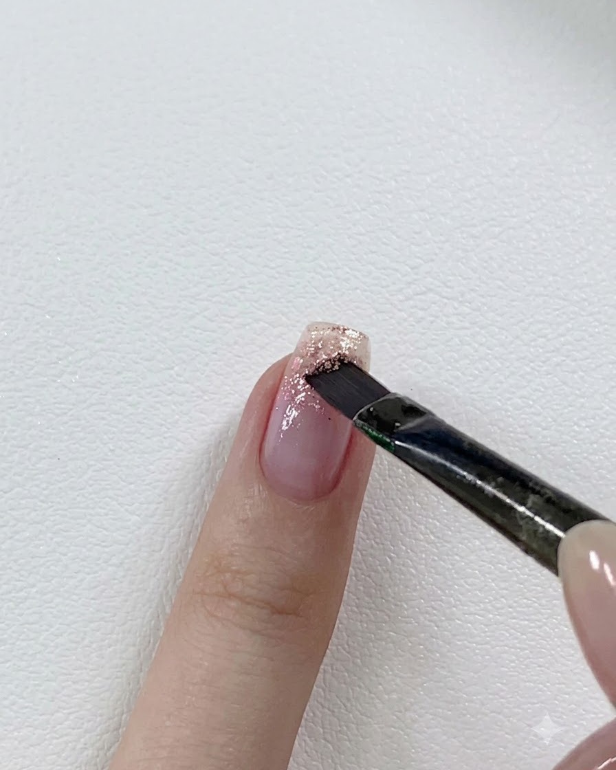 Un pinceau à ongles superposant un gel pailleté rose plus foncé sur le bord libre dun ongle partiellement pailleté.