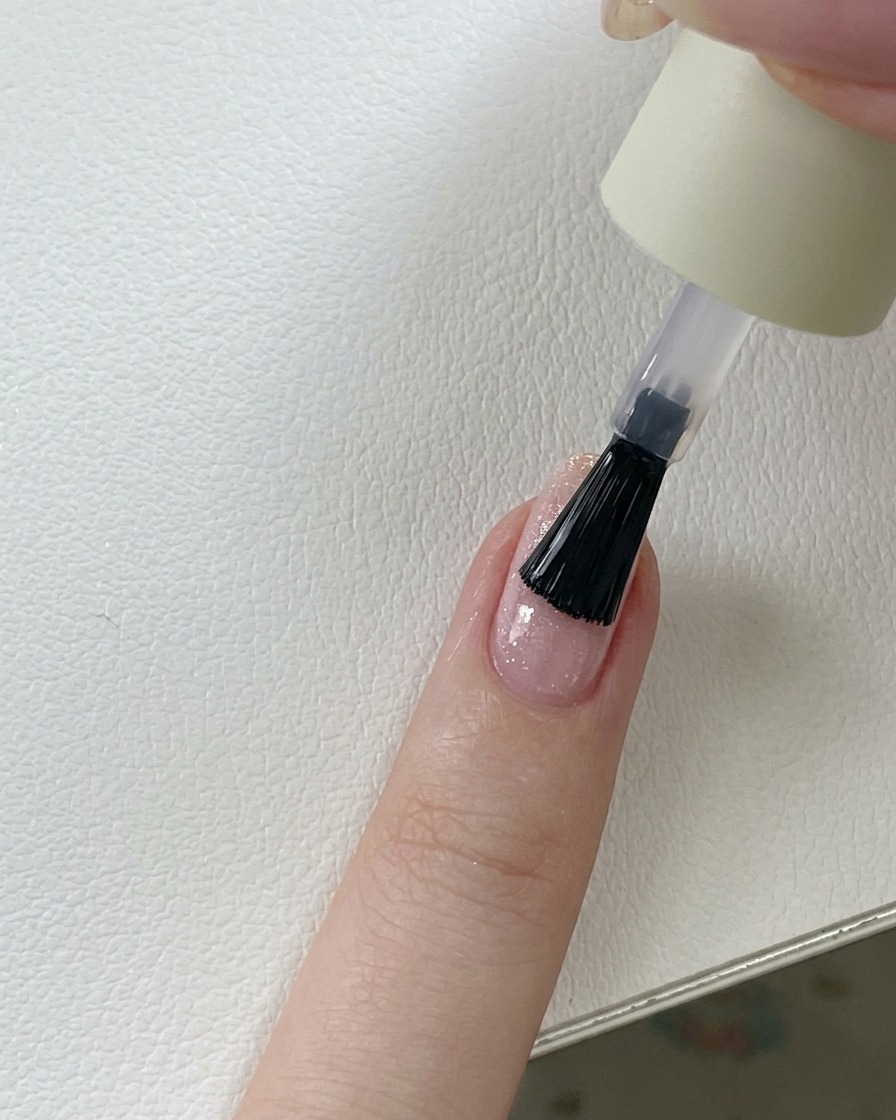 Un pinceau appliquant un top coat transparent et brillant sur un ongle en gel pailleté rose.