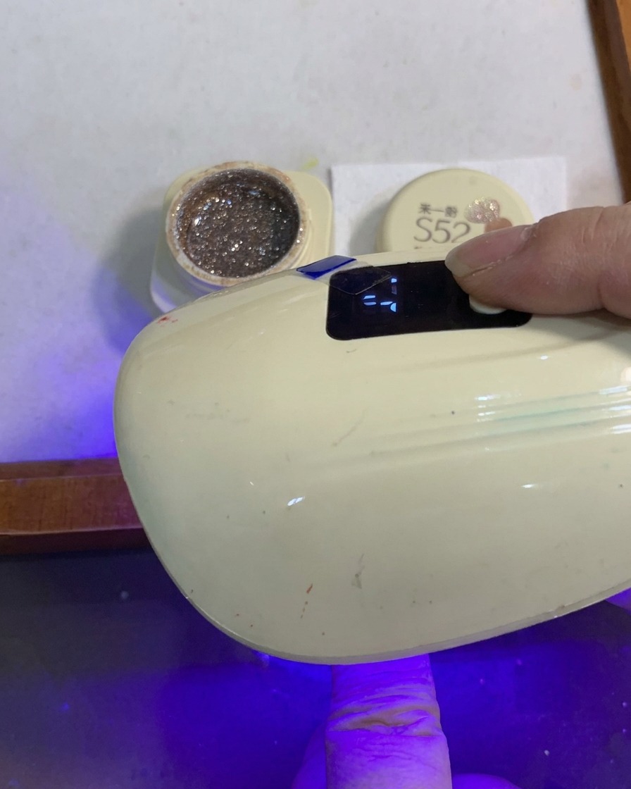 Un ongle avec un dégradé de paillettes en train de catalyser sous une petite lampe UV/LED portable.