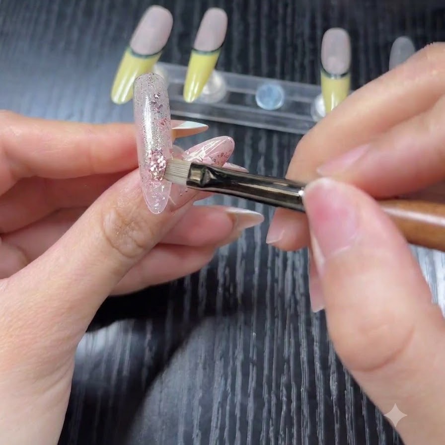 Un gros plan dun pinceau appliquant des paillettes holographiques roses et argentées sur une base transparente dun faux ongle.