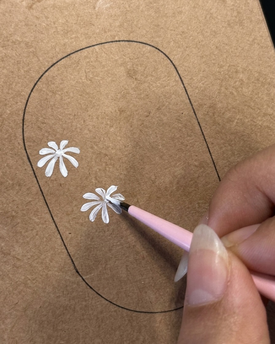 Un pinceau fin détaillant des pétales blancs sur un deuxième design de marguerite peint sur un modèle dentraînement marron.