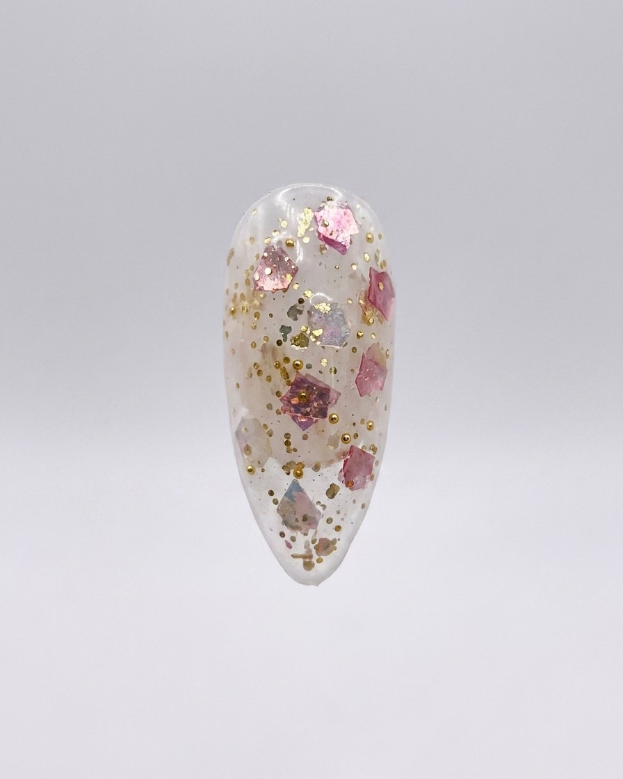 Un ongle press-on transparent parsemé de paillettes roses et dorées, isolé sur fond gris clair.