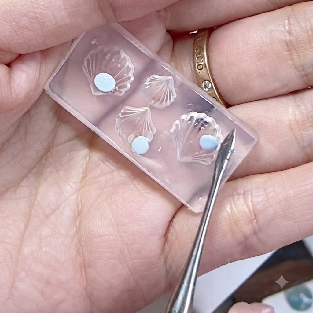 Un moule en silicone tenu dans une main avec des perles de gel bleu aplaties et un outil de modelage à proximité.