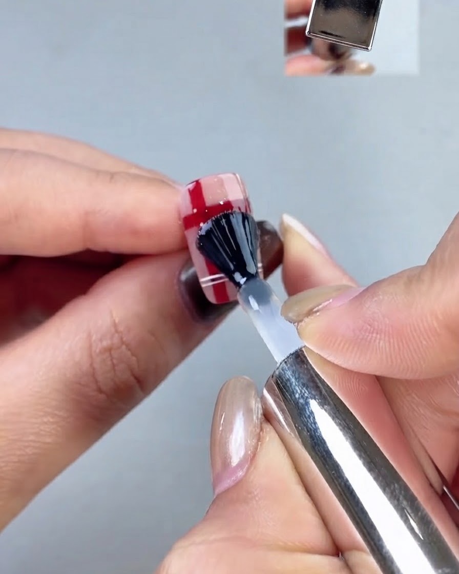 Un pinceau à ongle au capuchon noir appliquant une couche de finition transparente sur un motif dongle à carreaux.