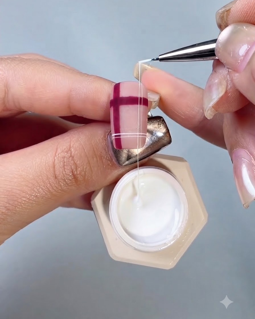 Un pinceau fin de nail art dessinant de fines lignes blanches avec du gel de soie sur un motif écossais rouge sur un ongle.