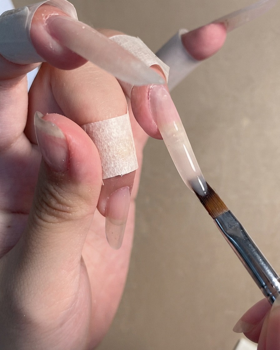 Un pinceau à ongle appliquant un gel polish nude diaphane sur une extension dongle stiletto longue.