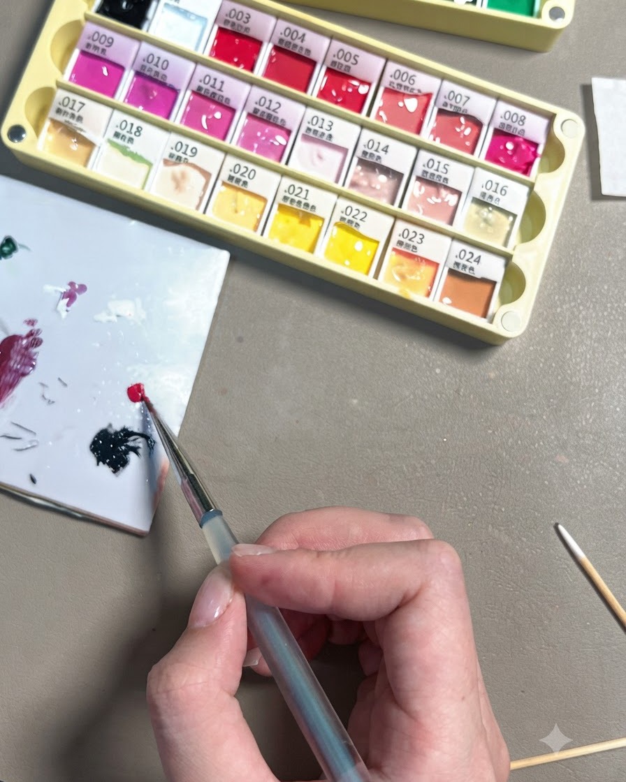 Une main utilisant un pinceau de détail fin pour déposer une petite goutte de gel rouge rose vif sur une palette de mélange en céramique blanche.