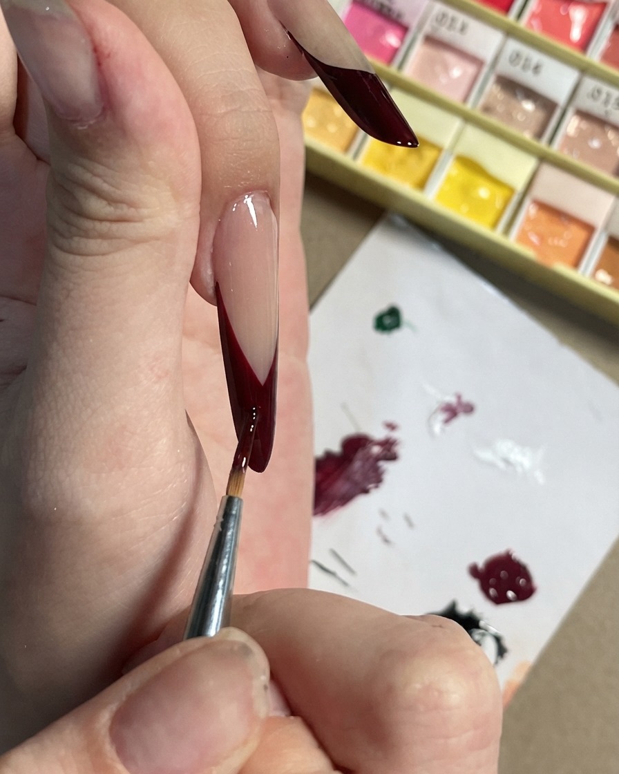 Un pinceau fin apposant de petits accents prune foncé et des formes de pétales sur une French tip noire.