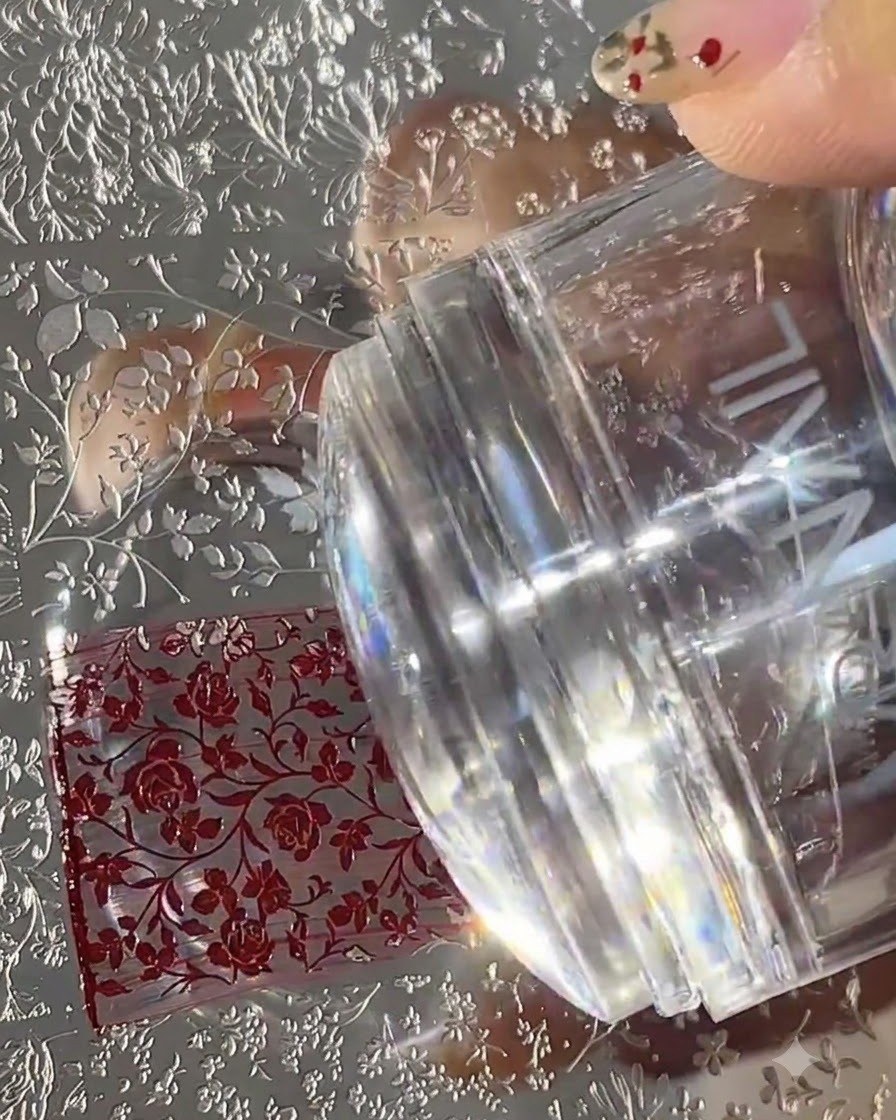 Un tampon en silicone transparent appuyé sur une plaque de stamping en métal remplie de motifs floraux rouges.