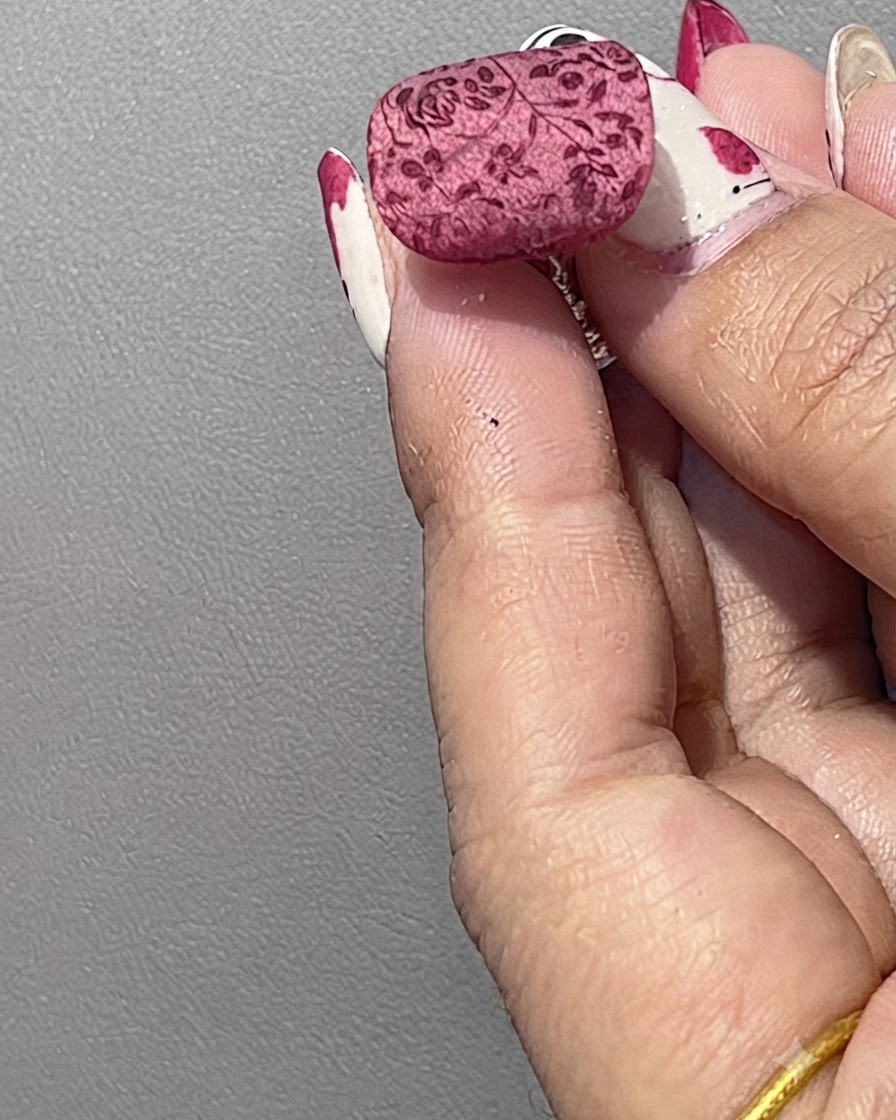 Un ongle manucuré avec un motif floral rouge estampé, légèrement recouvert de poudre de cristal blanche à laide dune spatule.