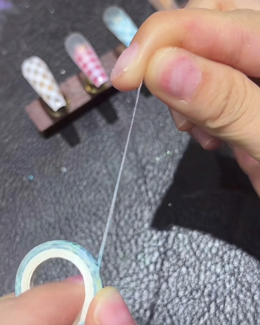 Une paire de brucelles plaçant du ruban de striping fin en diagonale sur un ongle de couleur nude.