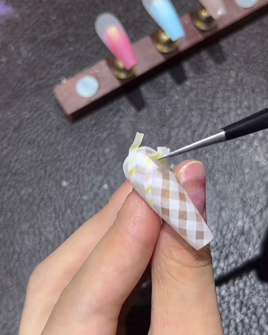 Un pinceau de détail fin appliquant une couche de finition brillante sur un ongle au motif écossais marron terminé.
