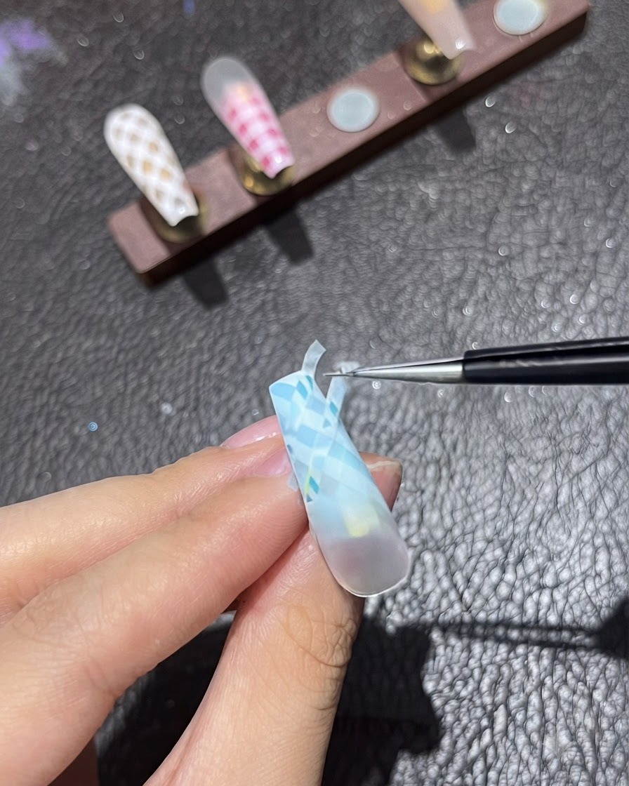 Un outil à ongles fin positionnant une bande de ruban dans la direction diagonale inversée sur un ongle aux tons bleus.